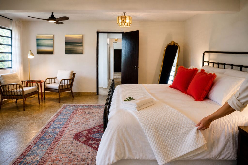 Airbnb bedroom being prepared for guests with neatly made bed, folded towels, and cozy seating area in a warm tropical interior