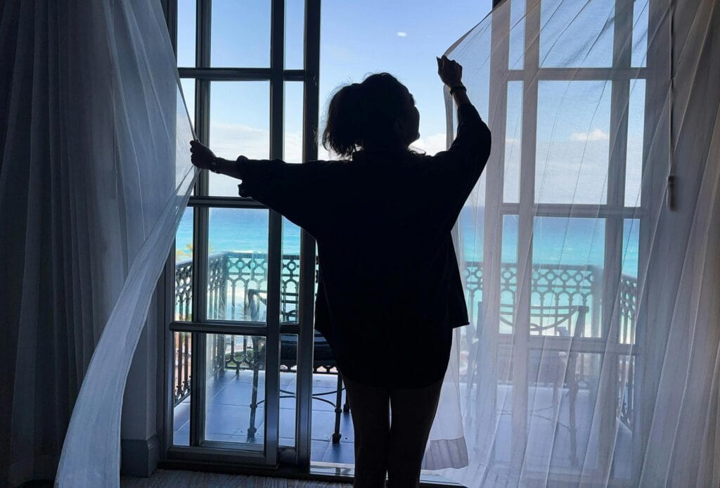 Guest opening curtains to ocean view from Airbnb balcony, representing the decision moment after clicking a listing but before booking