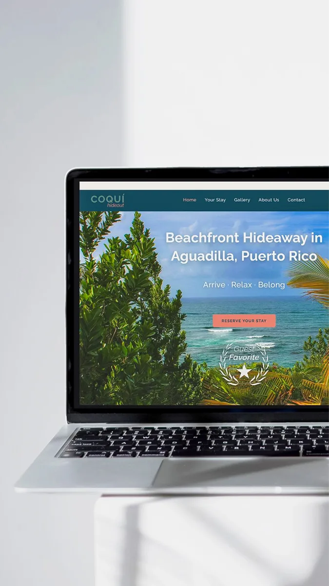 Laptop showing the live Coquí Hideout vacation rental website with tropical beach design, coral booking button, and ocean background, placed on a white table in natural light.